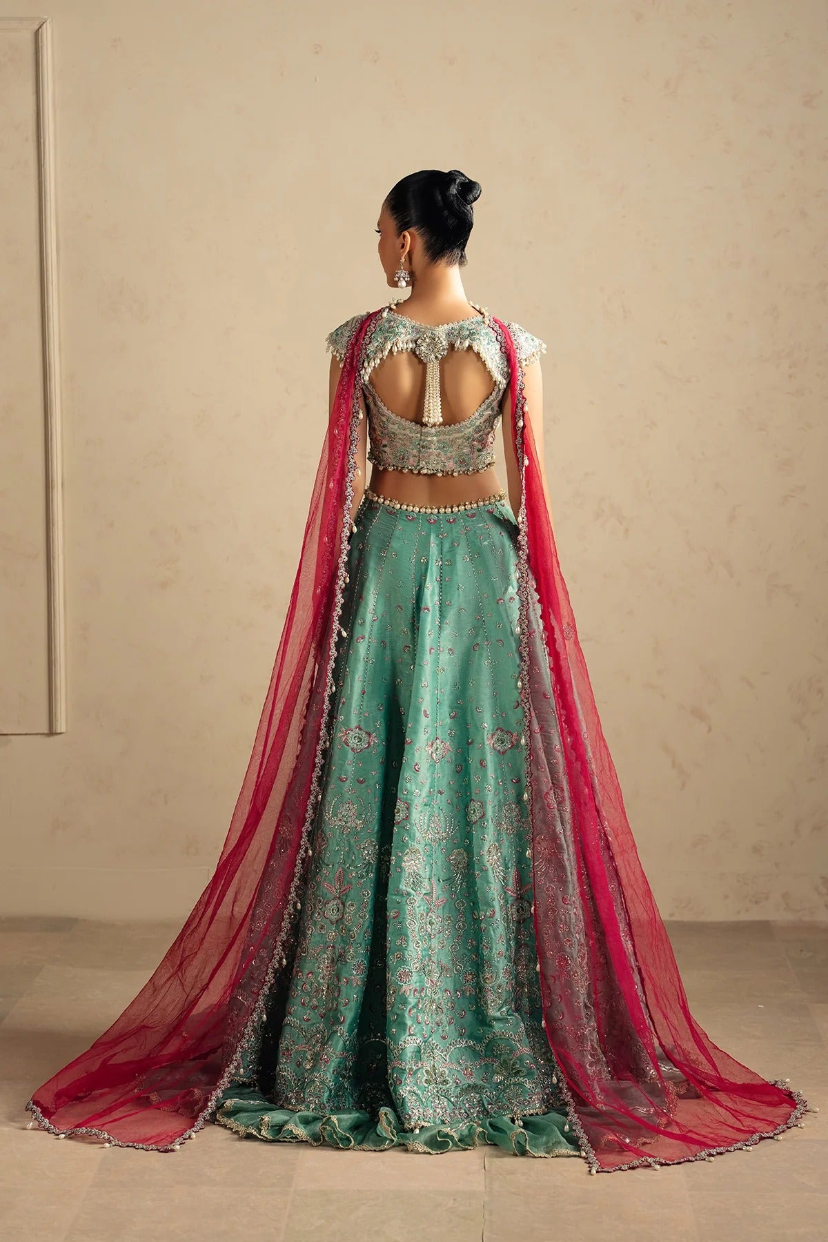 A woman stands with her back to the camera wearing an ornate teal-green embroidered lehenga choli with a long pink dupatta, set against a neutral beige wall.