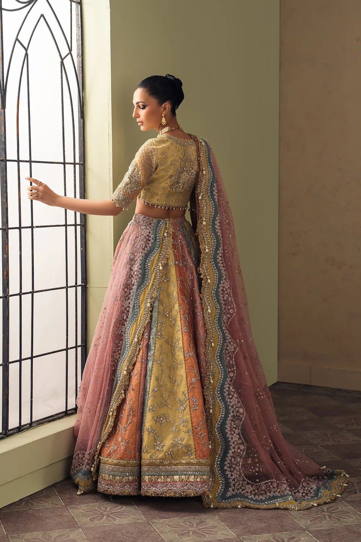 A woman wearing an ornate traditional Indian lehenga choli with a dusty pink dupatta, standing by a tall arched window in a beige room.