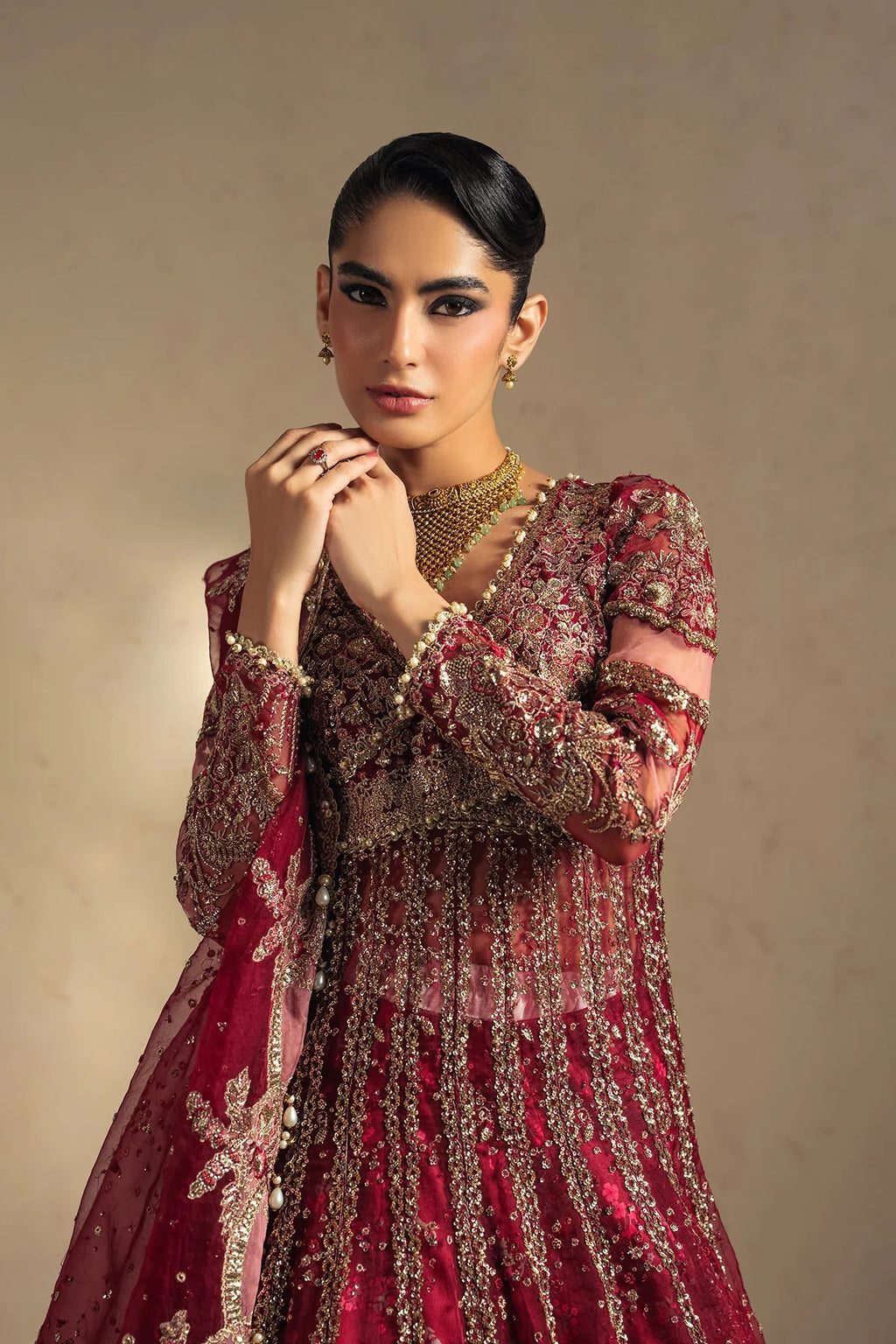 A woman modeling a heavily embellished maroon and gold traditional embroidered dress with a matching sheer dupatta, set against a neutral beige studio background.