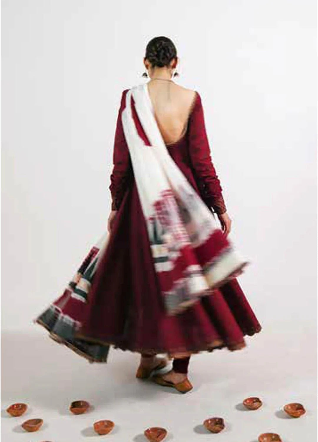 A woman viewed from behind wearing a deep maroon long-sleeved dress with a wide open back and a white dupatta featuring red and black patterns, set against a plain light gray background with small decorative clay pots on the floor.