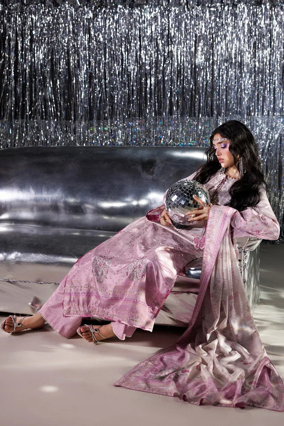 A woman in a pink embroidered traditional outfit sits on a metallic sofa holding a reflective silver orb in front of a silver tinsel backdrop.