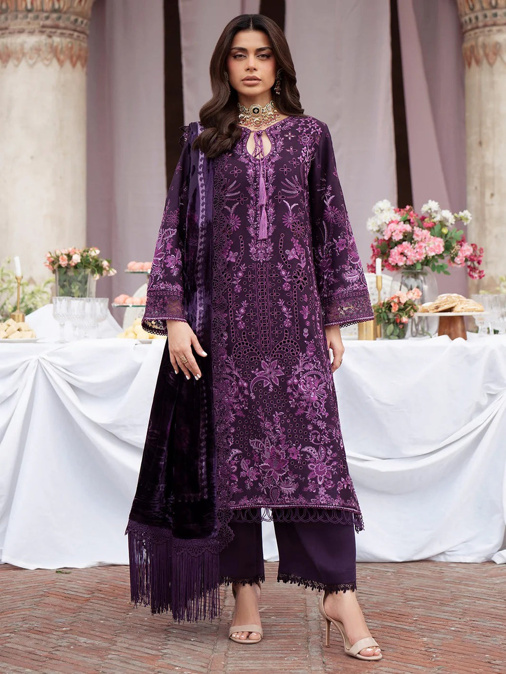 A woman models a traditional three-piece purple embroidered kurta set with matching pants and dupatta in an elegant outdoor event setting.
