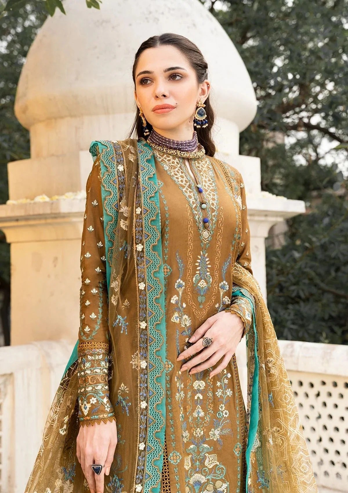 A woman wearing a traditional embroidered brown and teal kurta set with a matching dupatta, standing outdoors in front of a stone architectural structure.