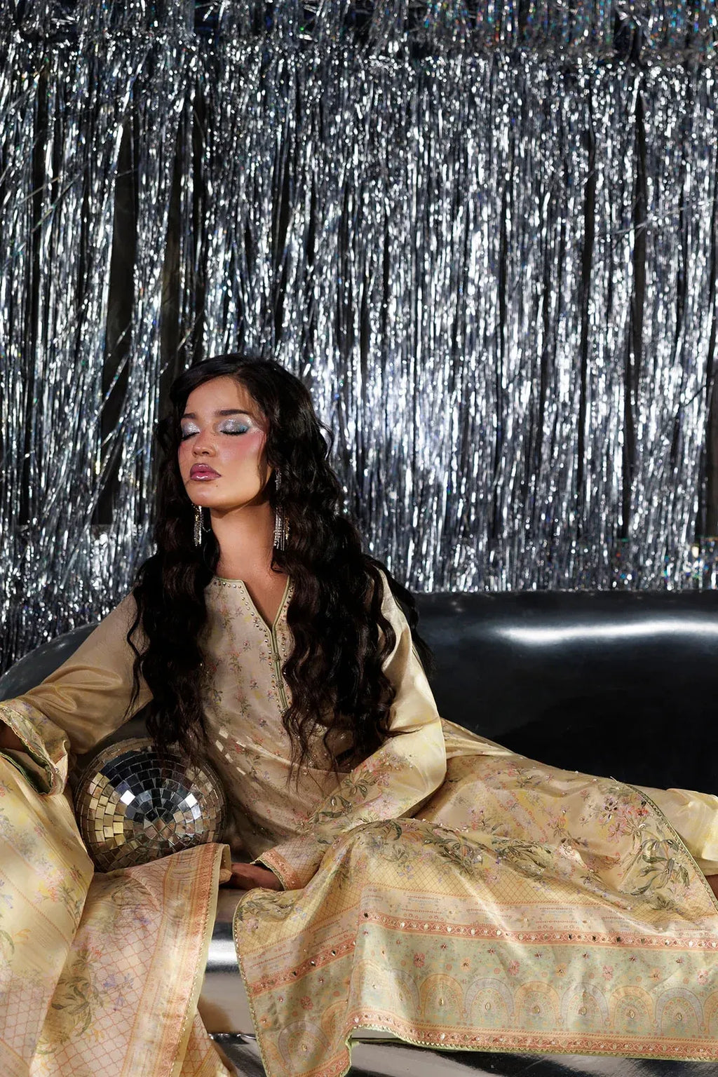 A woman with long dark wavy hair sits on a black leather couch in front of a silver tinsel curtain backdrop, wearing a beige embroidered traditional outfit with a matching dupatta and holding a small disco ball.
