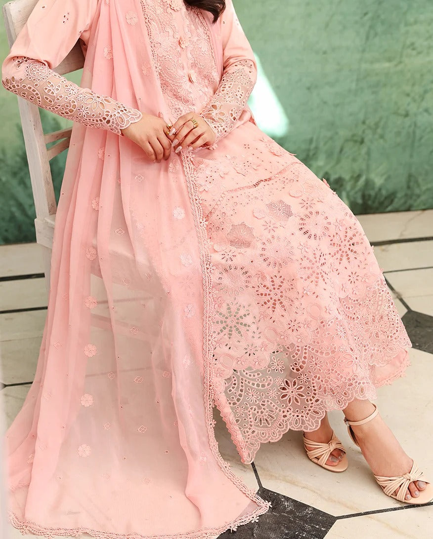 A woman wearing a light pink traditional outfit with a long embroidered kurta, matching sheer dupatta, and lace-trimmed skirt, seated on a white chair against a green textured wall.