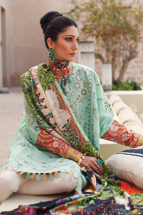 A woman seated outdoors wearing a mint green embroidered traditional outfit with a multicolored printed dupatta and patterned sleeves, styled with turquoise jewelry.