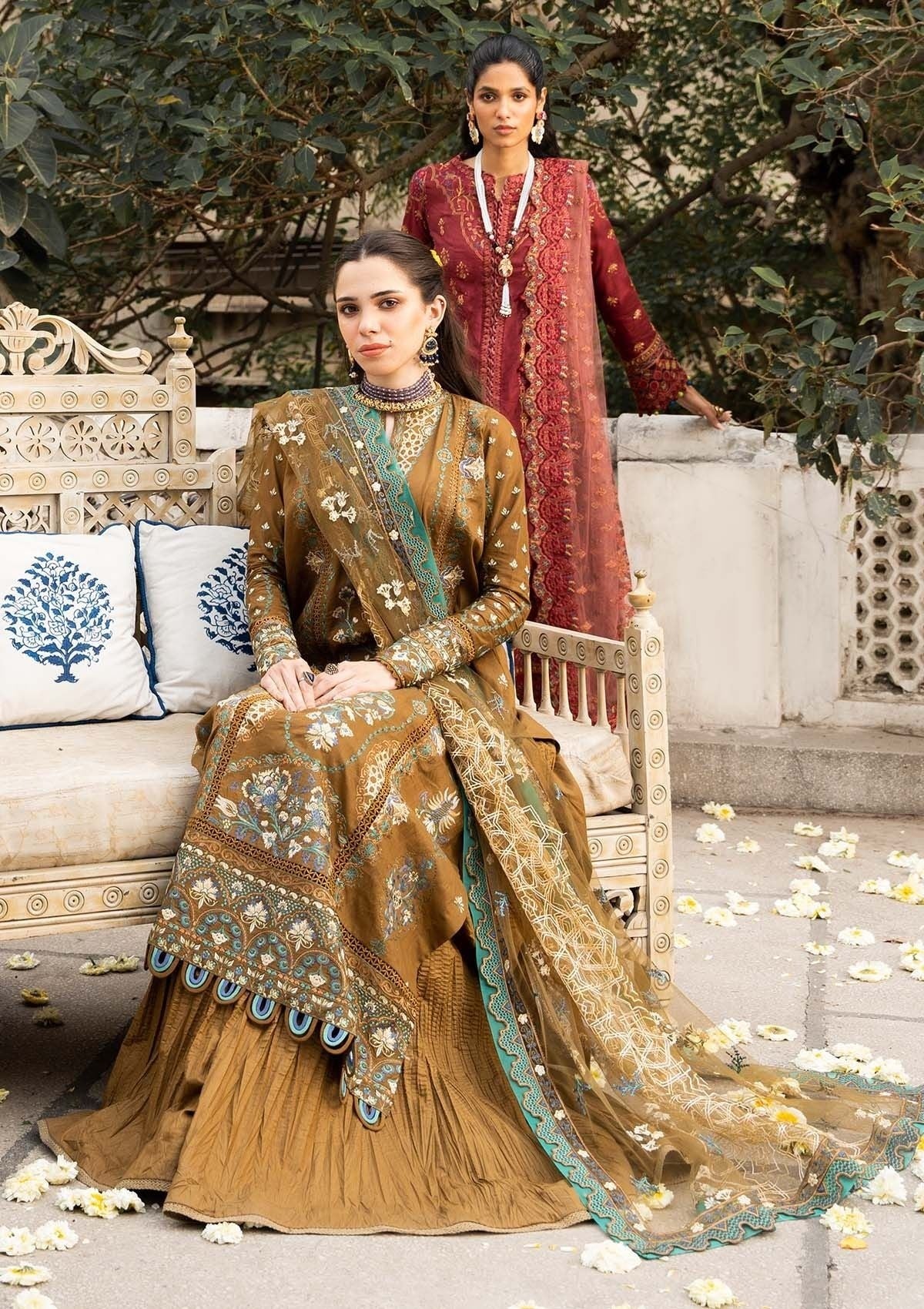 Two women in traditional embroidered dresses sitting on a decorative bench with floral arrangements.