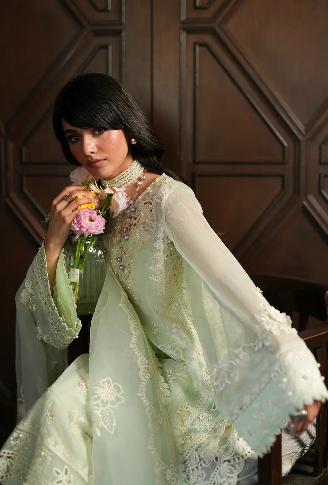 A woman wearing a light mint green embroidered traditional outfit with sheer long sleeves and lace detailing, holding a small bouquet of pink flowers in front of a dark wood paneled background.