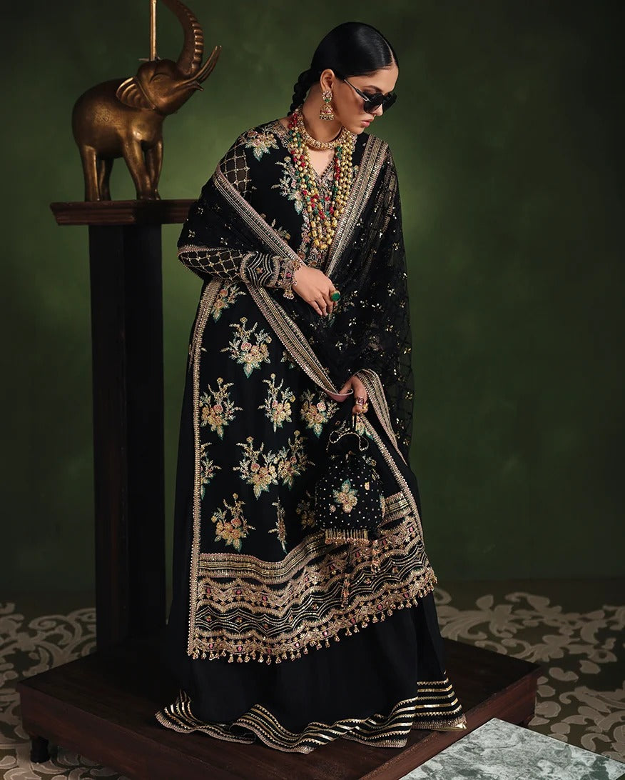 A woman models a black embroidered traditional outfit with a matching dupatta, featuring intricate gold, beige, and green floral embroidery, set against a dark green studio background with a decorative elephant statue.
