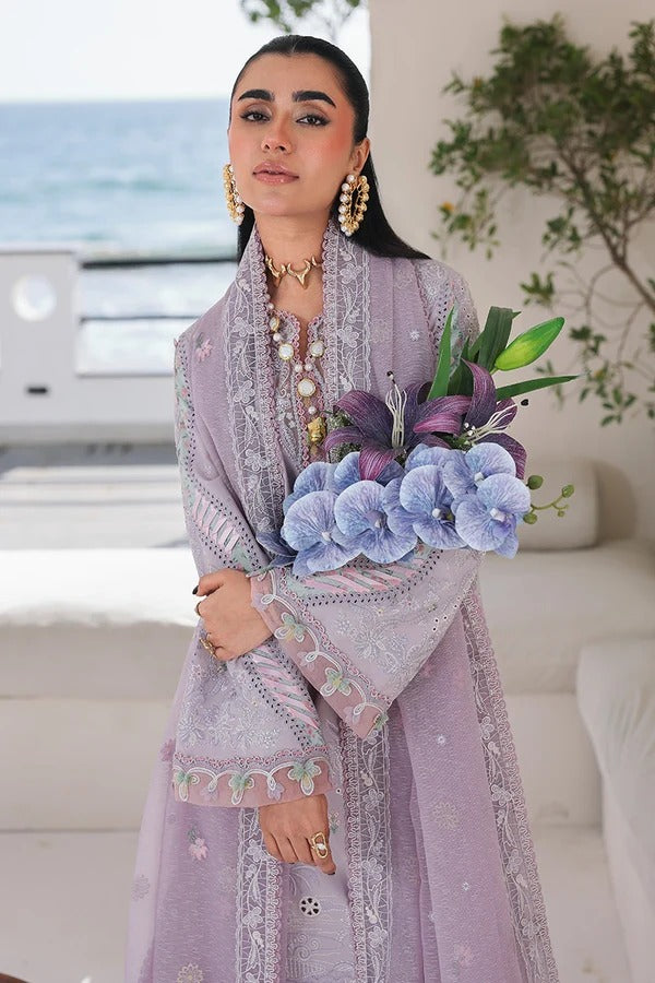 A woman wearing a lavender traditional embroidered outfit holds a bouquet of light blue and purple flowers in a bright indoor setting with a blurred ocean and plant background.