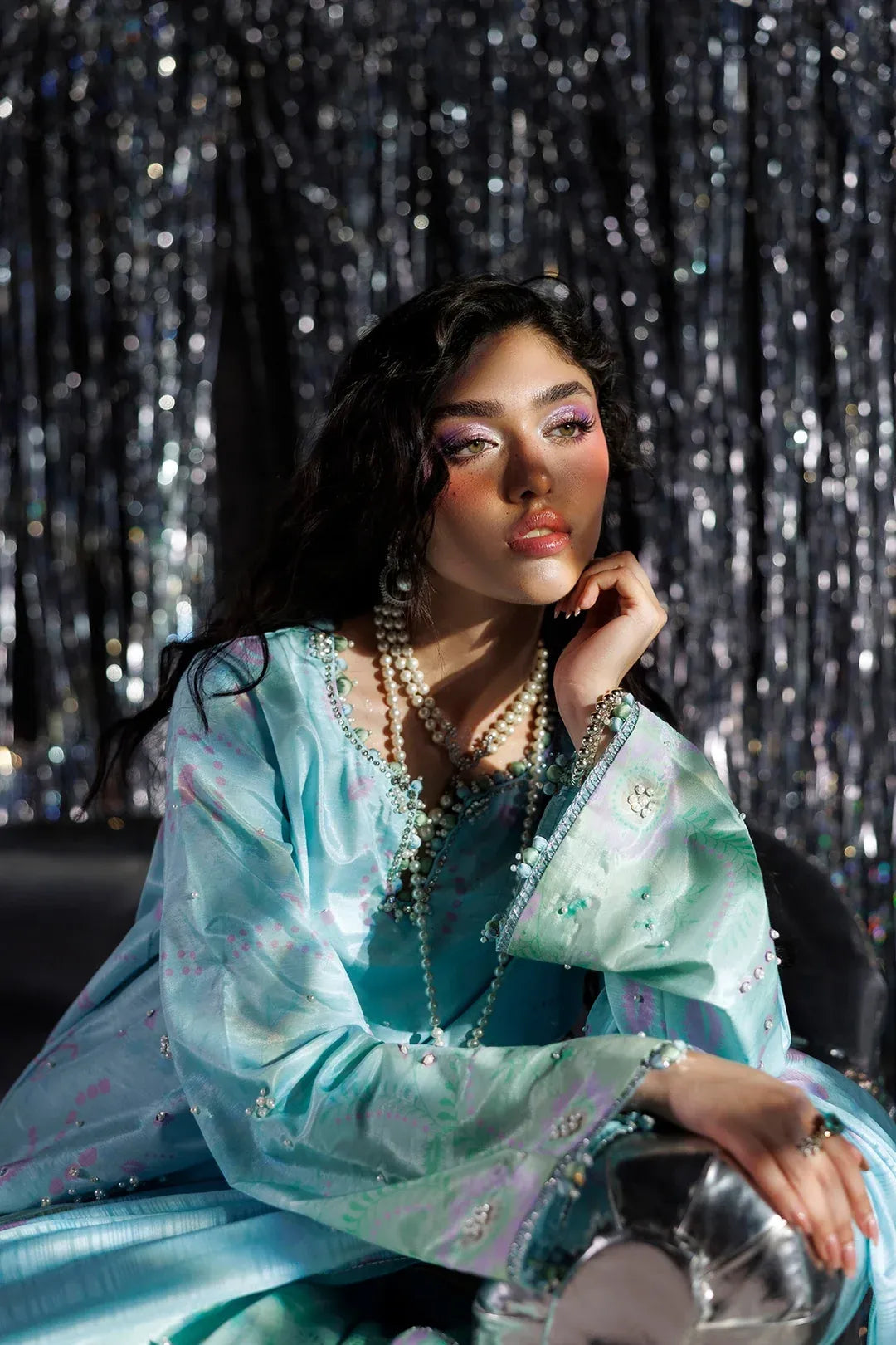 A woman posing in a light blue embroidered traditional outfit with silver embellishments, layered with multiple pearl necklaces, set against a shimmering silver tinsel backdrop.