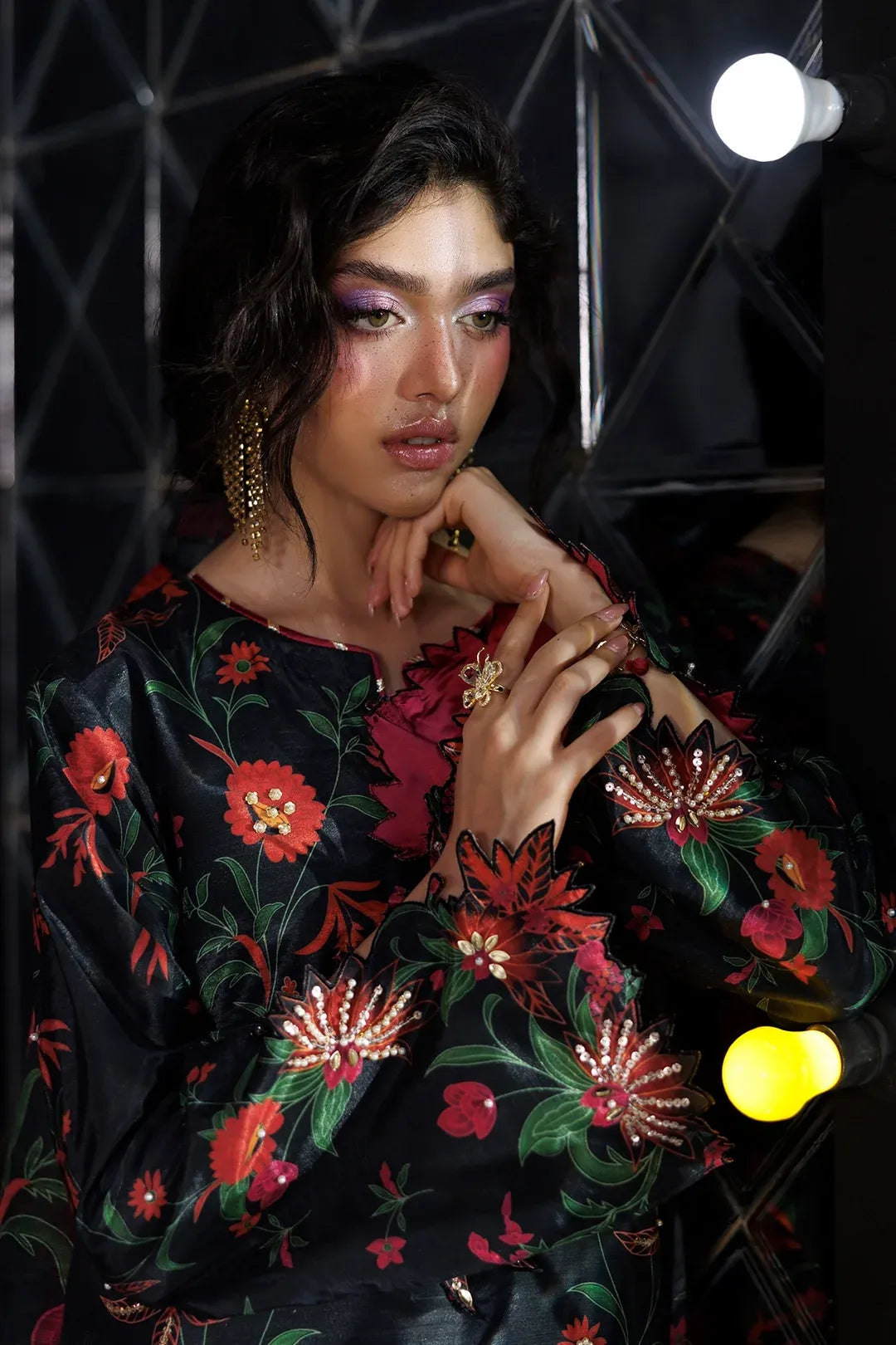 A model wearing a black satin long-sleeve top with a vibrant red, pink, and green floral print, posed against a dark studio background with lighting fixtures.