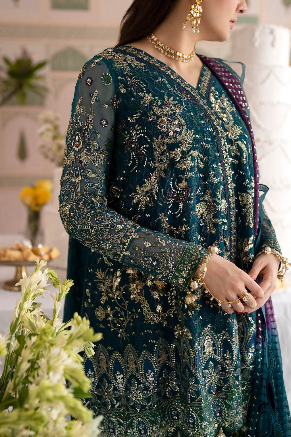 A woman wearing an ornate teal blue and gold embroidered traditional outfit with a purple dupatta, photographed indoors with decorative plants and warm lighting.
