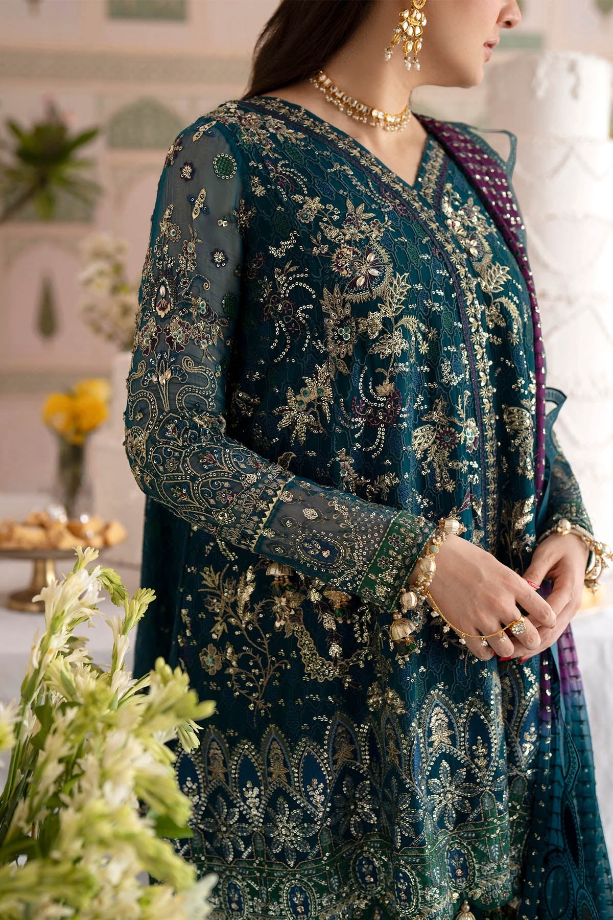 A woman wearing an ornate teal blue and gold embroidered traditional outfit with a purple dupatta, photographed indoors with decorative plants and warm lighting.