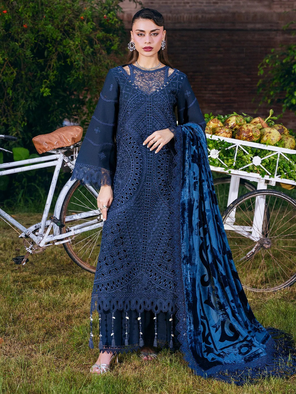 A woman models a dark navy blue embroidered ethnic outfit with a matching dupatta, standing outdoors on grass with a bicycle and greenery in the background.