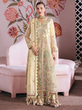 A woman models a traditional embroidered Anarkali suit in pale yellow and mint green with pink floral details, standing in front of a pink sofa and a pink wall decorated with floral and flamingo motifs.