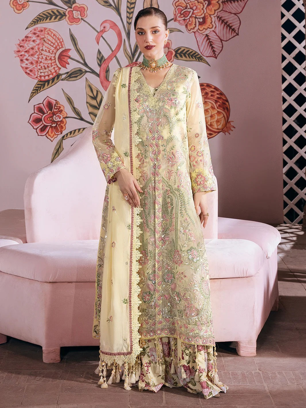 A woman models a traditional embroidered Anarkali suit in pale yellow and mint green with pink floral details, standing in front of a pink sofa and a pink wall decorated with floral and flamingo motifs.