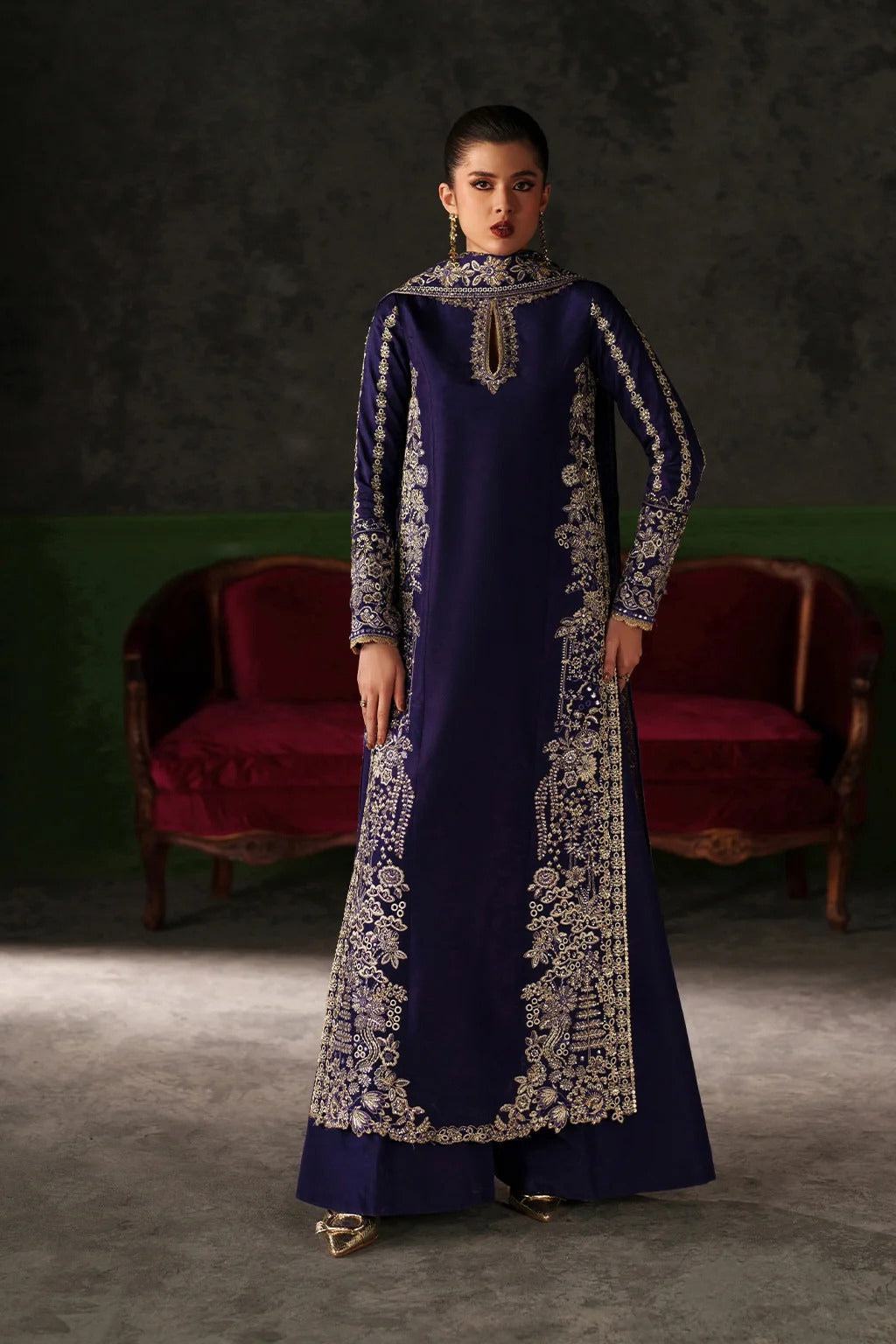 A woman stands in a studio wearing a long navy blue traditional dress with ornate silver embroidery and a high embroidered collar. Behind her are two red velvet armchairs against a dark textured wall.