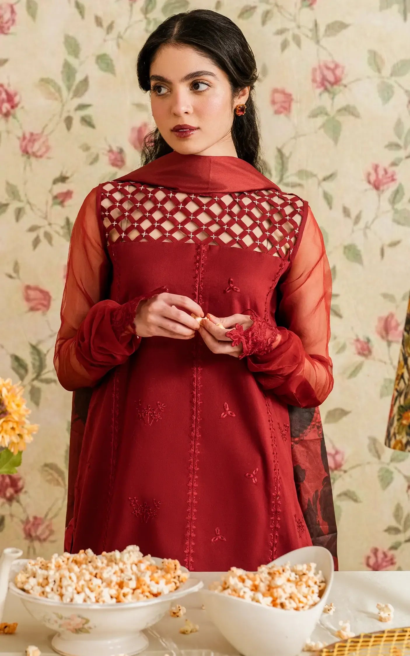 A woman wearing a deep red embroidered traditional outfit with sheer sleeves and a matching headscarf, standing in front of a floral wallpaper background with bowls of popcorn on a table in the foreground.