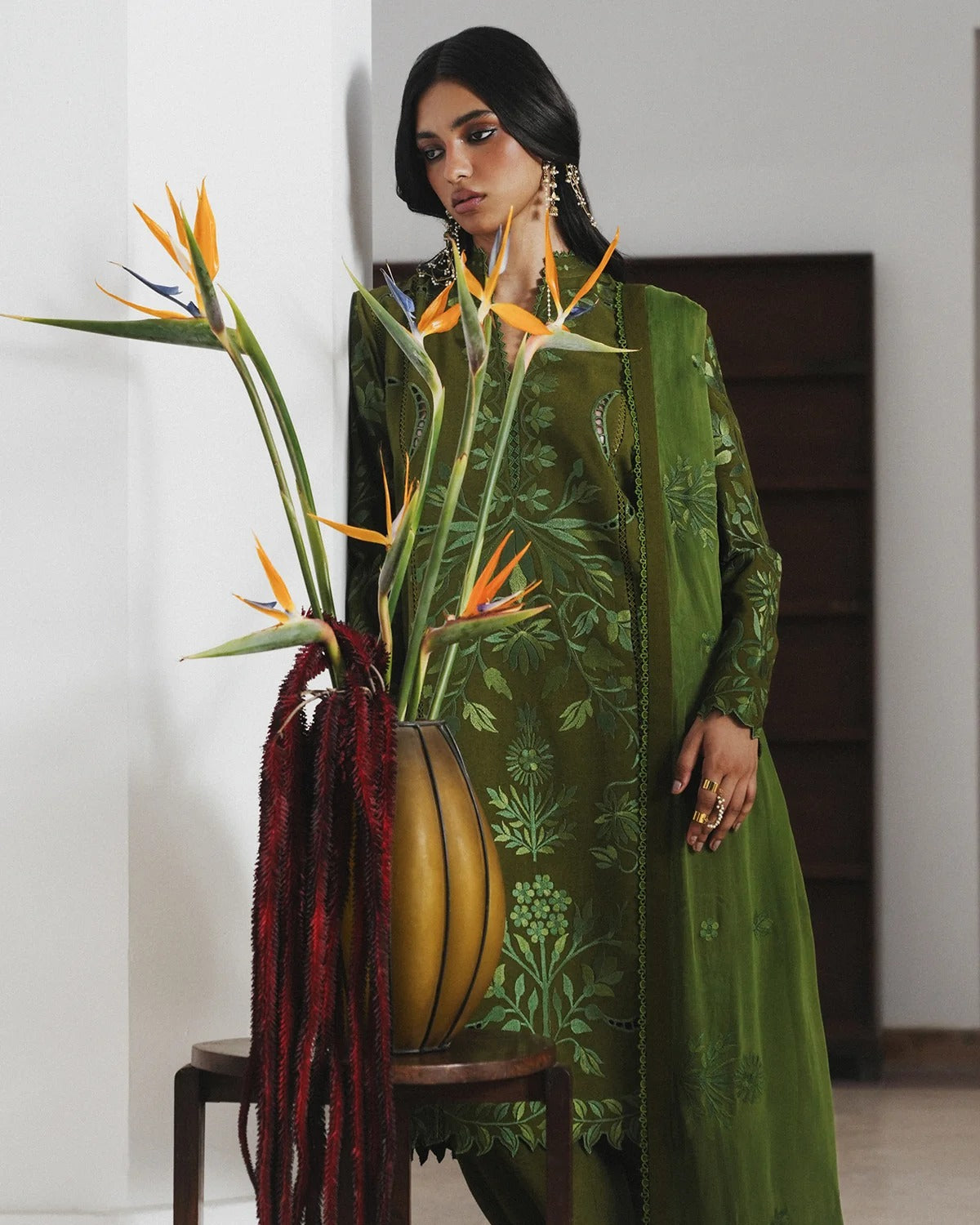 A woman models an olive green embroidered traditional outfit with a matching dupatta, standing indoors next to a large vase filled with bird of paradise flowers.