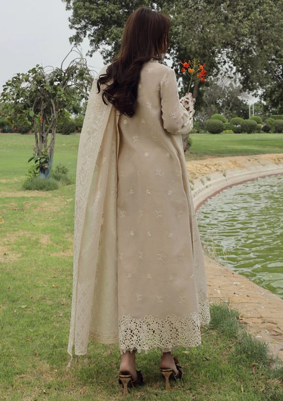 A woman stands outdoors by a pond wearing a long beige embroidered dress with a matching dupatta and black heeled sandals, holding a small bouquet of flowers.