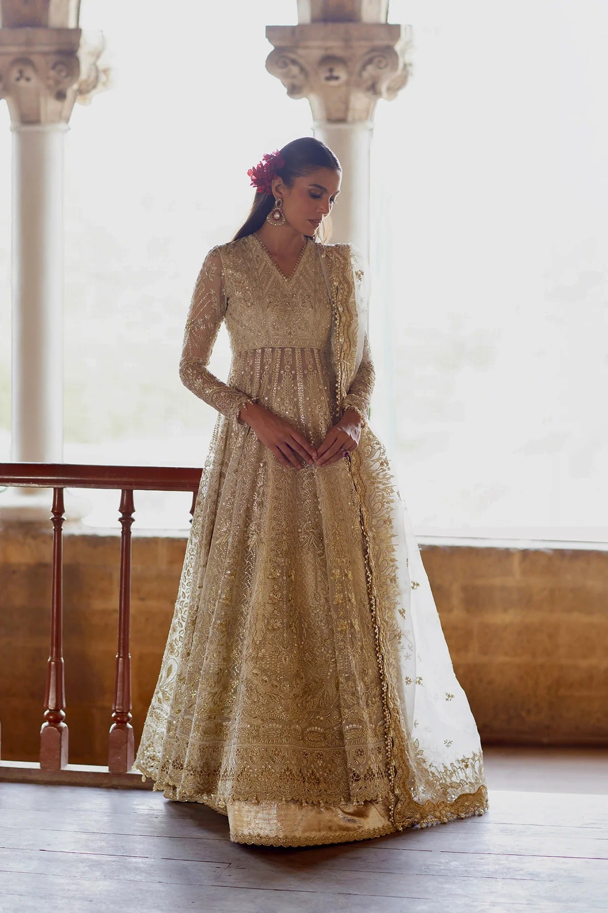 A woman wearing a heavily embroidered beige and gold traditional gown with a matching sheer dupatta, standing in a bright, classical hall with stone pillars.