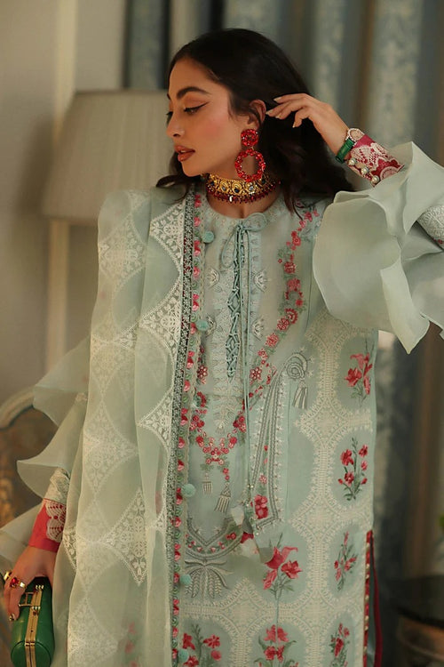 A woman wearing a pale mint green embroidered traditional outfit with floral patterns, lace details, and a sheer outer layer, styled with ornate red and gold jewelry in an indoor setting.