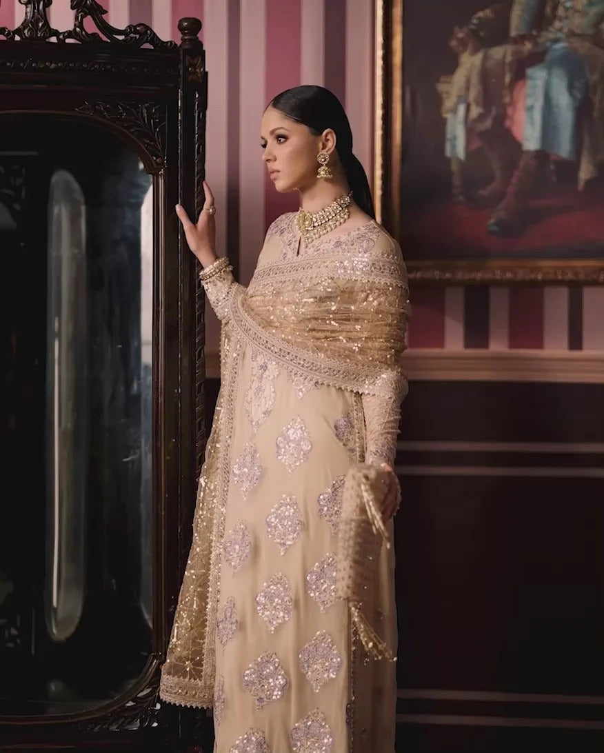 A woman wearing an ornate beige and gold heavily embellished traditional outfit with a matching dupatta, standing in a vintage room next to a large dark wooden mirror.