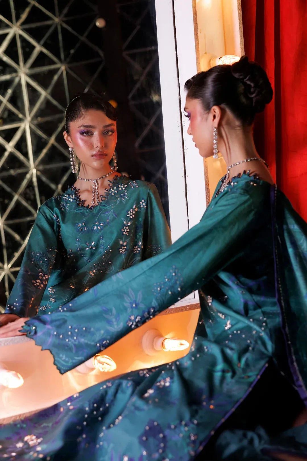 A woman in a teal embroidered traditional outfit poses in front of a mirror, showing both the front and side details of the dress. The background features a geometric black wall and a red curtain.