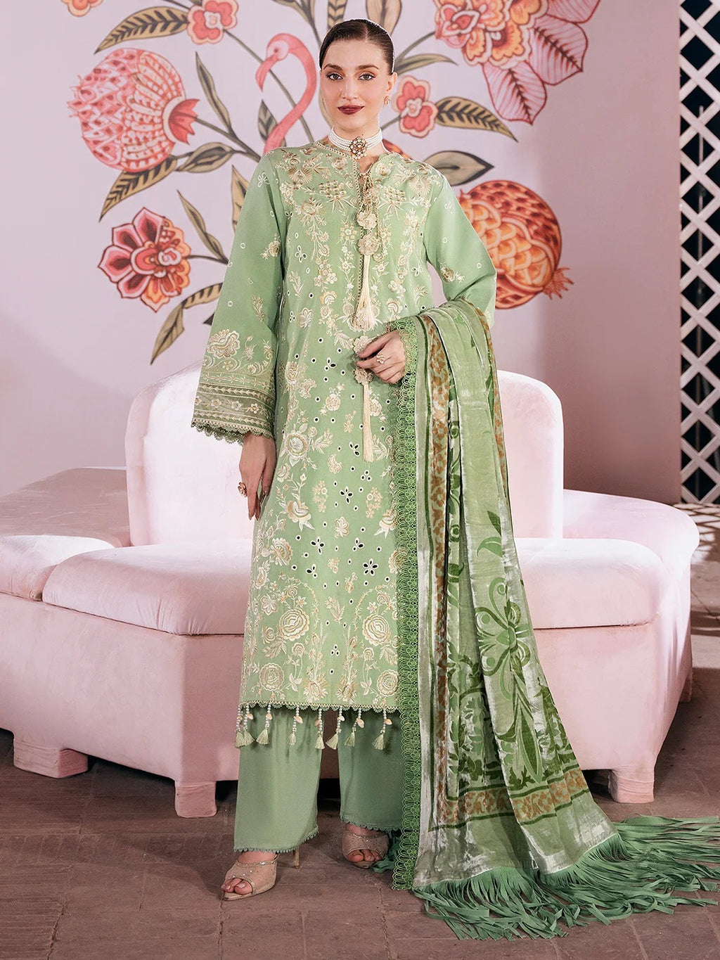 A woman models a light green three-piece traditional outfit with intricate gold embroidery, matching trousers, and a coordinating embroidered dupatta, posed in front of a pink sofa with a floral wall mural.