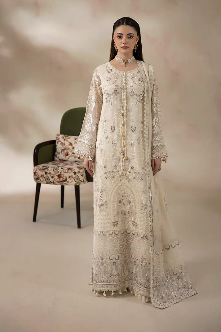 A woman models a traditional long cream-colored embroidered suit with matching dupatta in a studio setting. The outfit features intricate silver and gold embroidery, and is styled with a floral-patterned chair in the background.