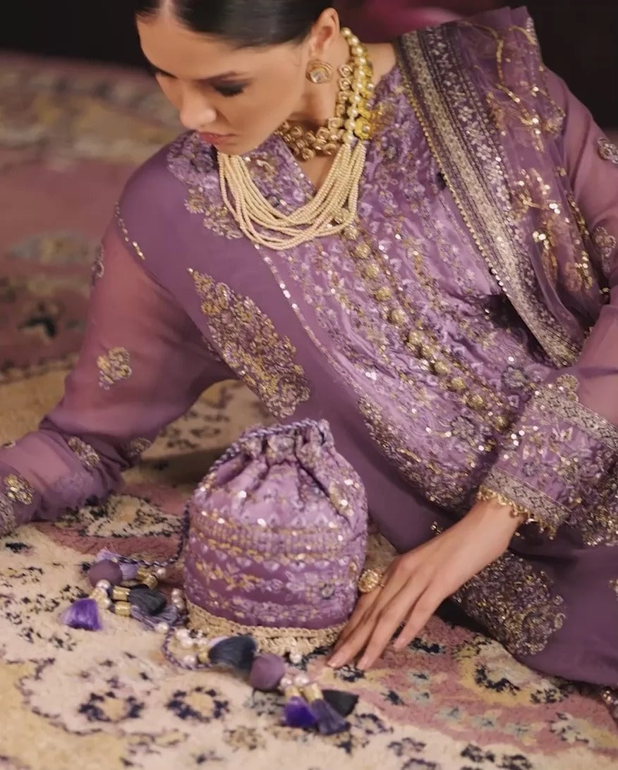 A woman wearing an ornate lavender purple and gold embroidered traditional outfit sits on a patterned rug, with a matching embroidered drawstring pouch placed in front of her.
