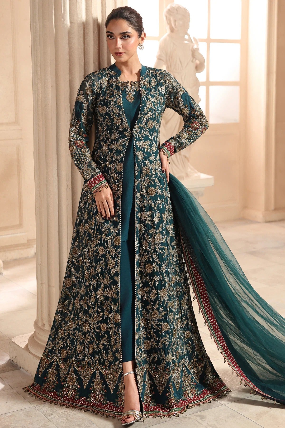 A woman models a long teal and gold embroidered Anarkali gown with a matching dupatta in an elegant indoor setting with classical columns and a statue.