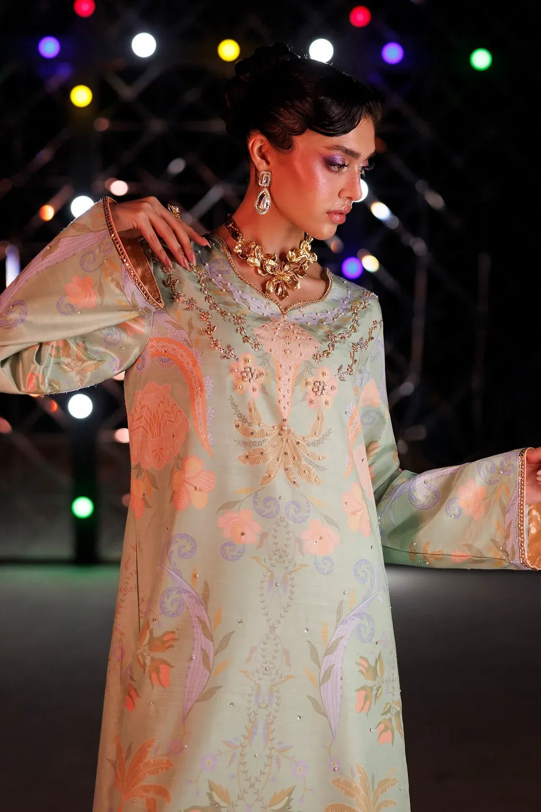A model wearing a light sage green traditional kurta with intricate peach, pink, and gold floral embroidery, accessorized with a gold necklace and earrings, set against a dark background with colorful stage lights.