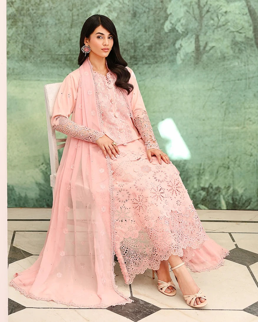 A woman models a light pink traditional outfit consisting of a heavily embroidered kurta, matching skirt, and a sheer dupatta, seated on a white chair against a green forest-patterned backdrop.