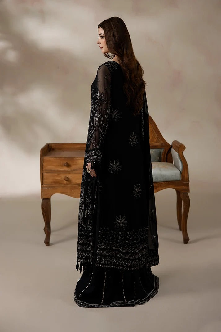 A woman stands in profile wearing a long black embroidered traditional outfit with sheer sleeves and ornate silver-toned detailing, posed in front of a wooden chair and cabinet against a warm beige studio backdrop.