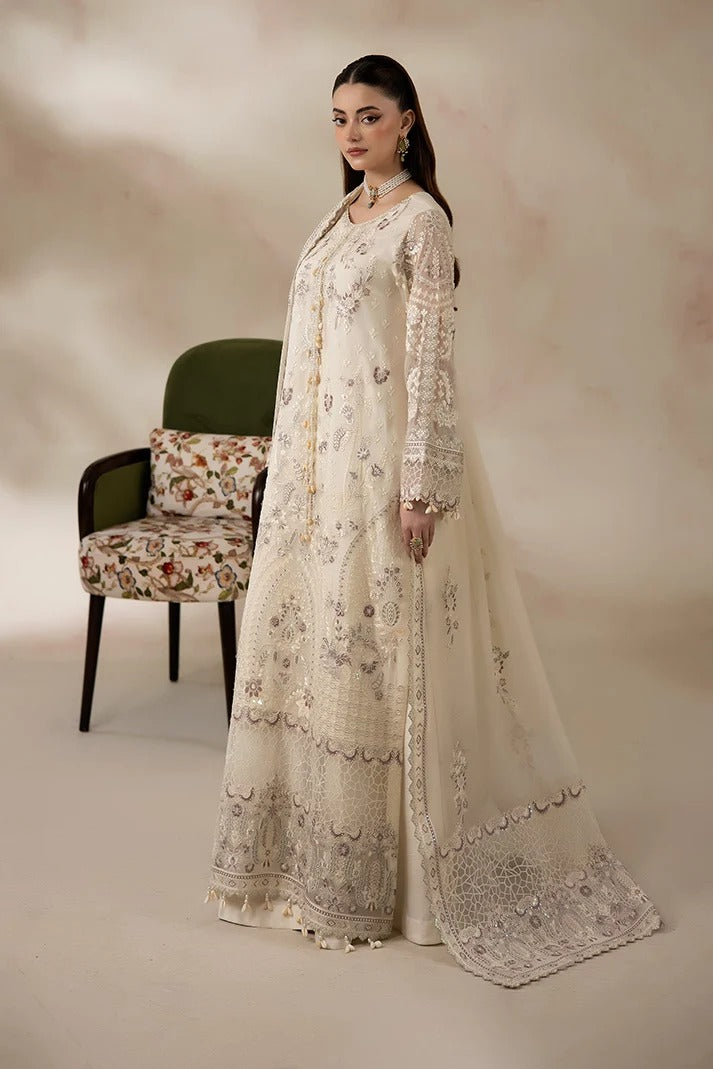 A woman models a traditional long cream-colored embroidered suit with a matching sheer dupatta, standing in a studio next to a green armchair.