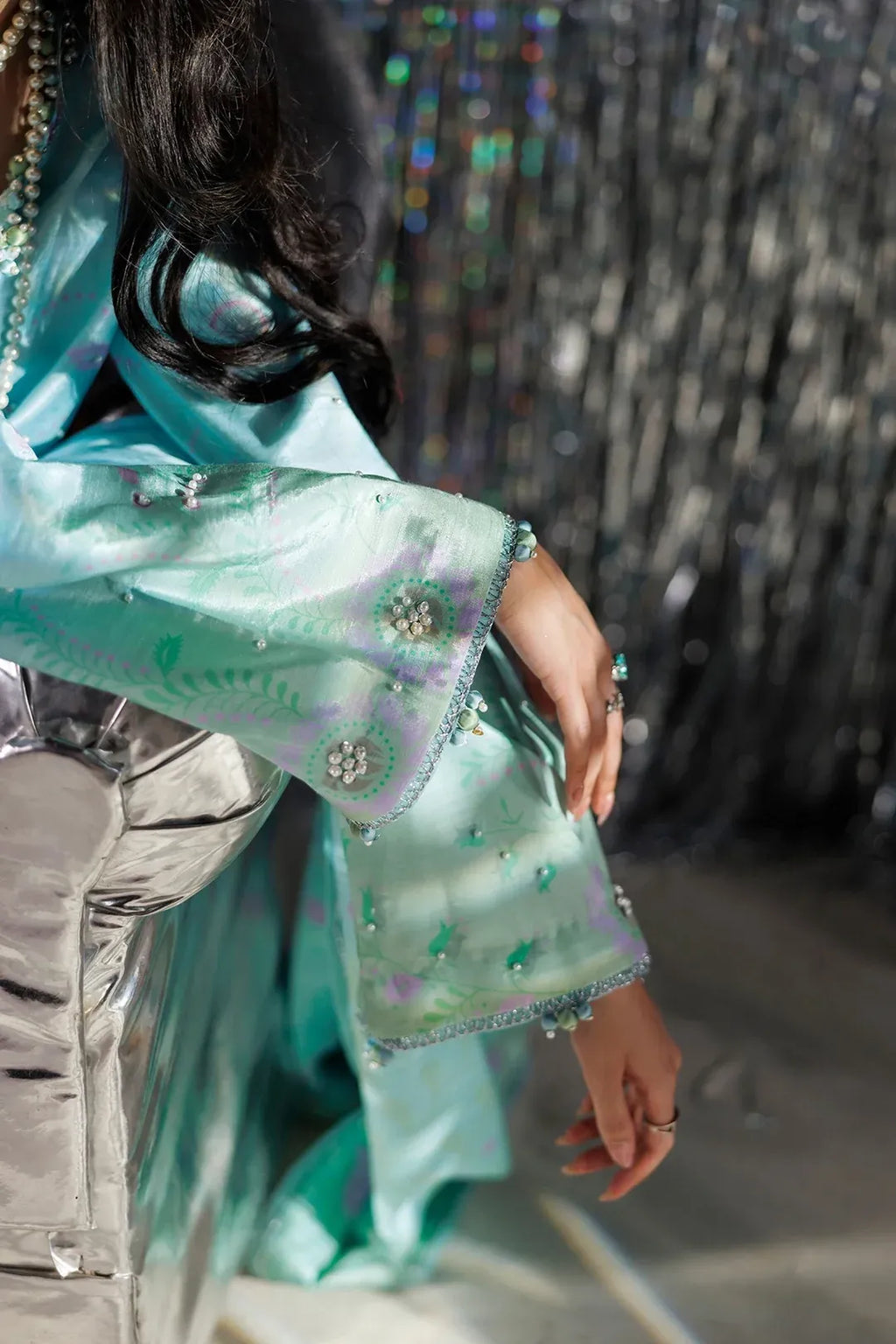 A close-up lifestyle shot of a model wearing a light turquoise green embroidered traditional garment with metallic silver sleeves, featuring intricate floral embroidery and beadwork.