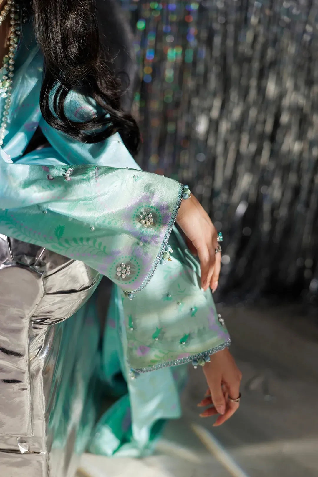 A close-up lifestyle shot of a model wearing a light turquoise green embroidered traditional garment with metallic silver sleeves, featuring intricate floral embroidery and beadwork.
