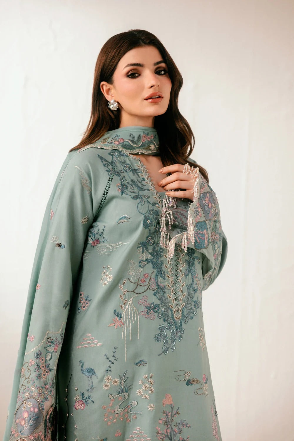 A woman modeling a sage green embroidered traditional outfit with floral and bird motifs, featuring a high collar, long sleeves, and delicate tassel details, set against a plain off-white background.