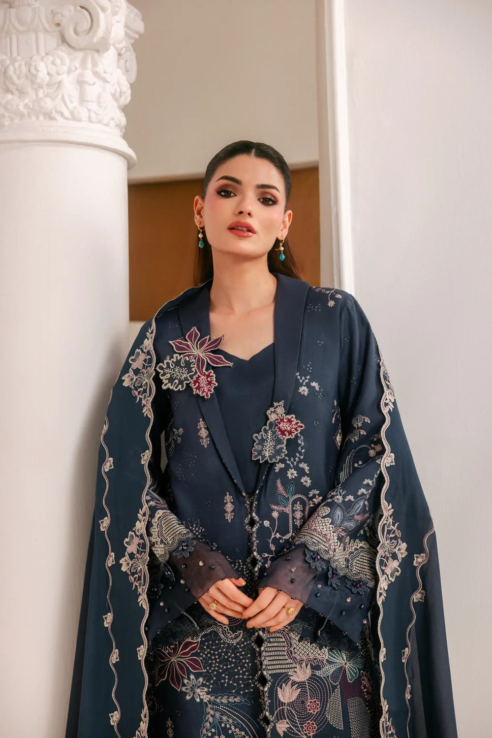 A woman models a dark navy blue traditional outfit with intricate floral embroidery in pink, white, and gold, set against a background featuring a white classical column.