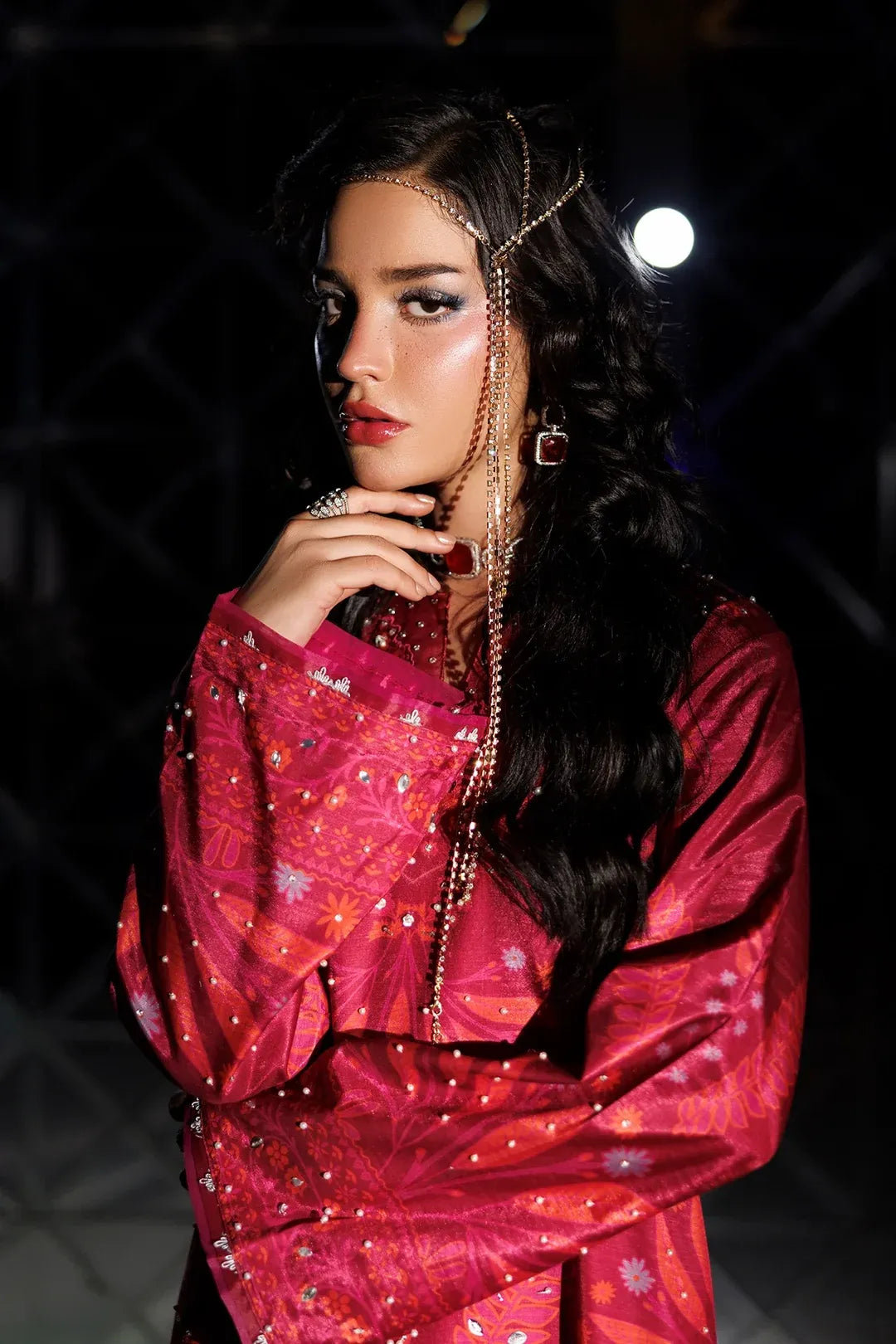 A woman with long dark wavy hair models a vibrant magenta-pink embroidered traditional outfit with gold star patterns, accessorized with a gold chain headpiece and matching earrings against a dark, moody background.