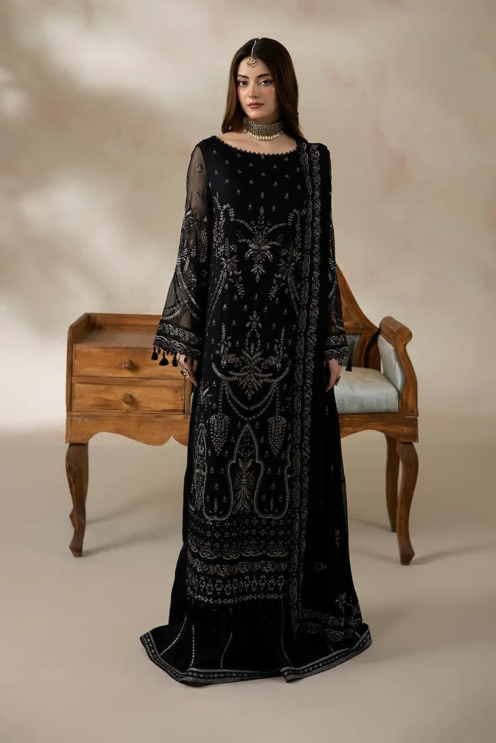 A woman models a long black embroidered dress with silver mirror-work detailing, standing in front of a wooden chair and cabinet against a beige studio backdrop.
