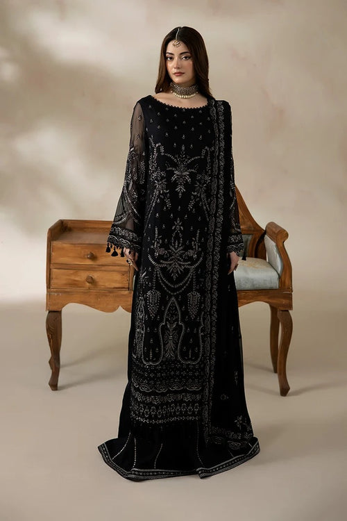 A woman models a long black embroidered dress with silver mirror-work detailing, standing in front of a wooden chair and cabinet against a beige studio backdrop.