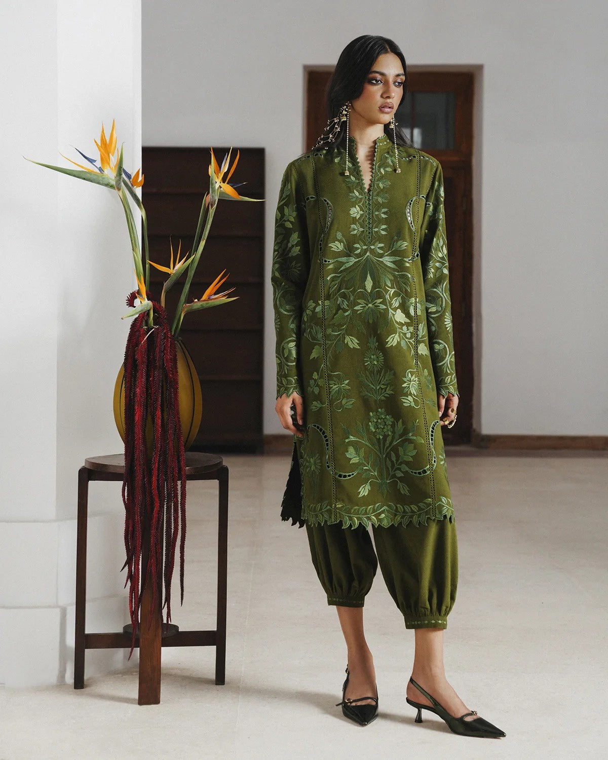 A woman models an olive green embroidered kurta and matching pants set in a minimalist indoor setting with a wooden stool and bird of paradise flowers.