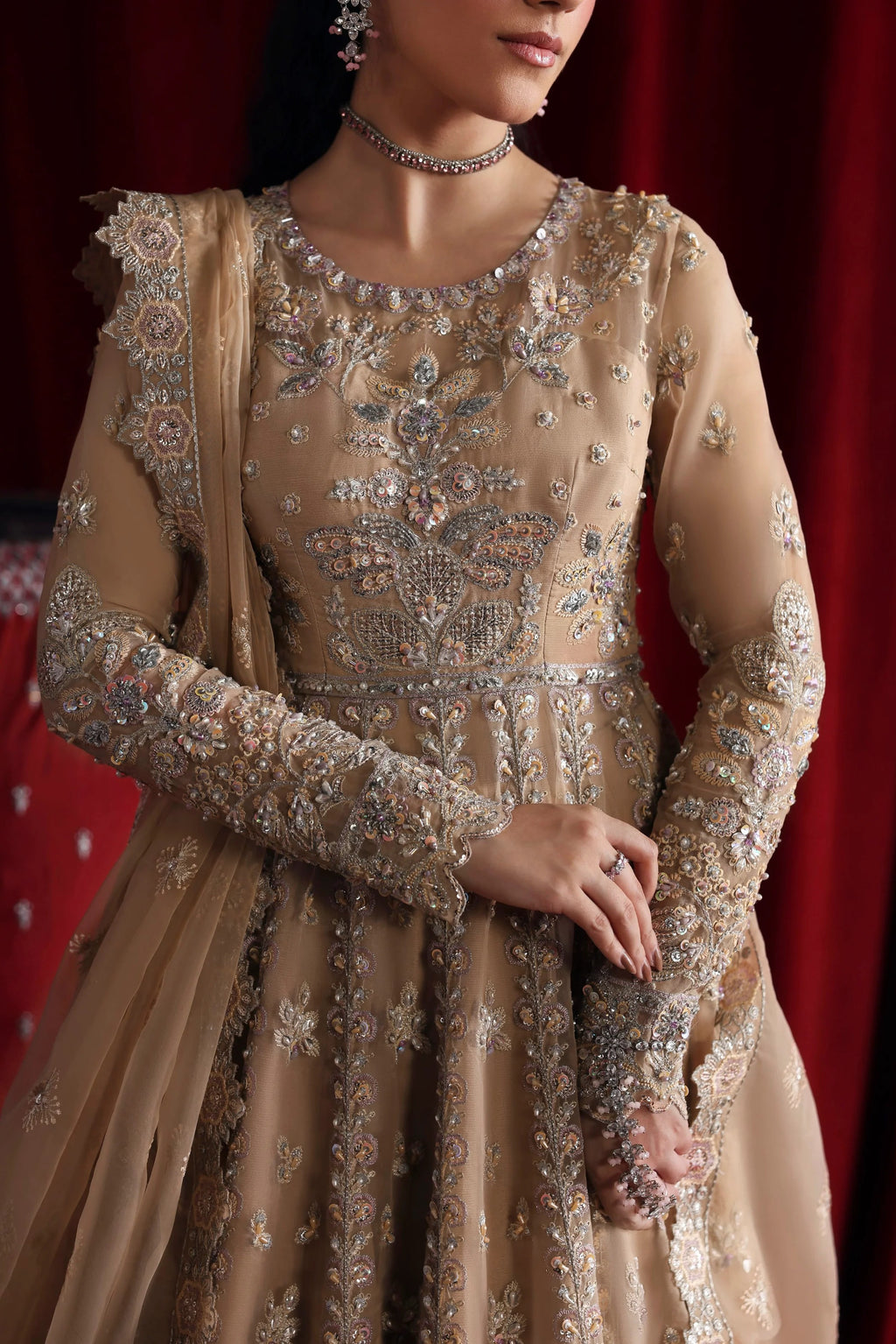 A woman models a heavily embellished beige and gold traditional gown with intricate silver embroidery, set against a dark red curtain backdrop.