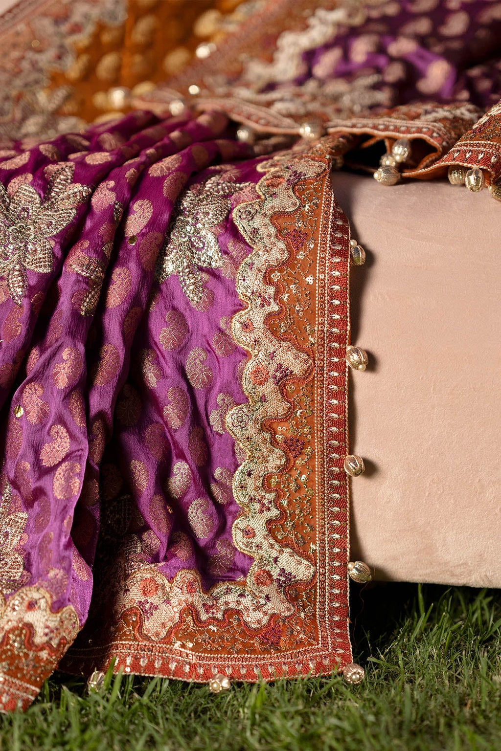 A close-up of an ornate purple and orange embroidered fabric draped over a beige cushion outdoors on grass, with detailed metallic embroidery and scalloped borders visible.