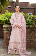 A woman stands outdoors wearing a light pink traditional outfit with gold embroidery, layered pearl necklaces, and a matching sheer dupatta draped over one shoulder. The background features a brick wall and green foliage.