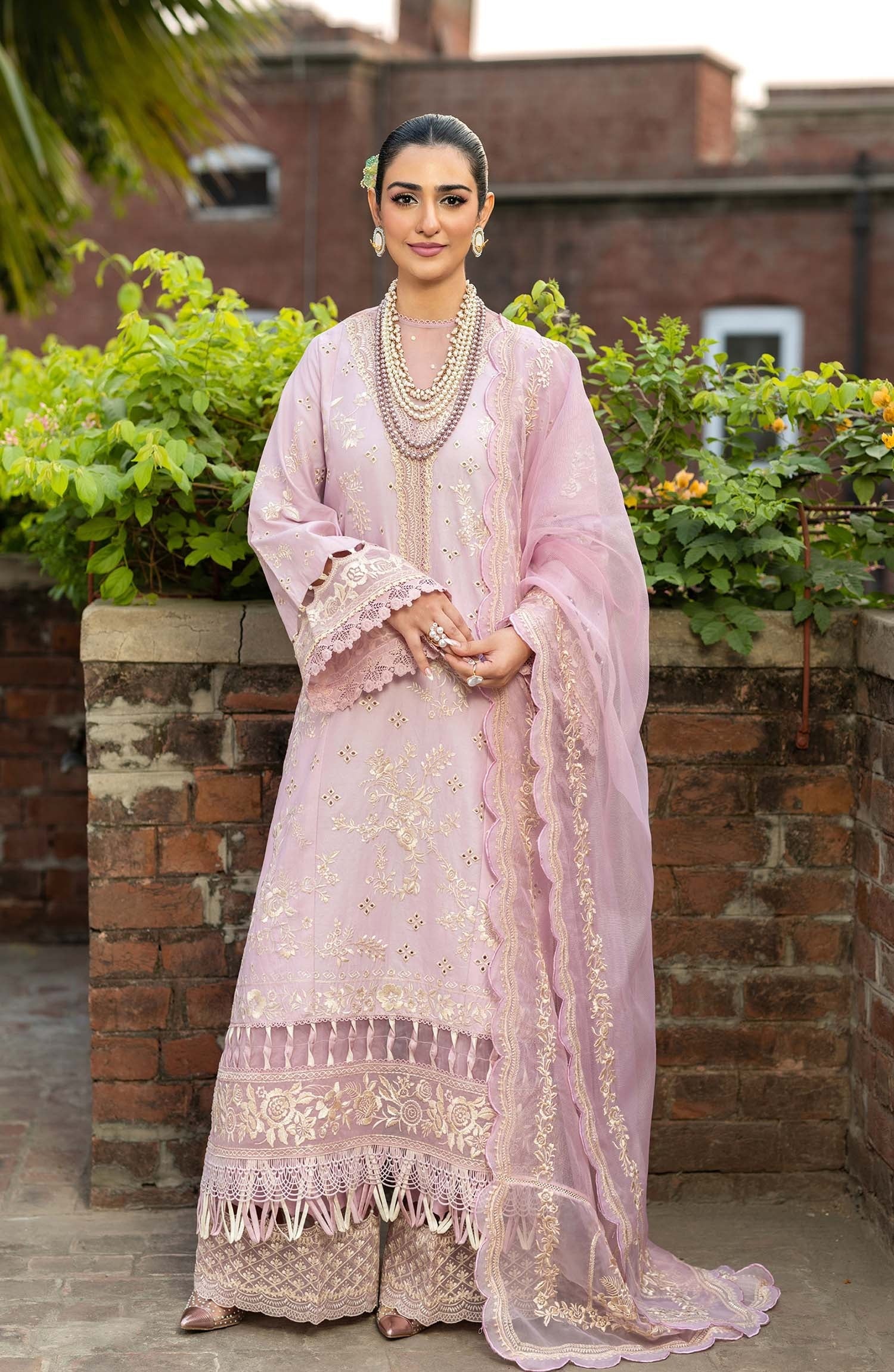 A woman stands outdoors wearing a light pink traditional outfit with gold embroidery, layered pearl necklaces, and a matching sheer dupatta draped over one shoulder. The background features a brick wall and green foliage.