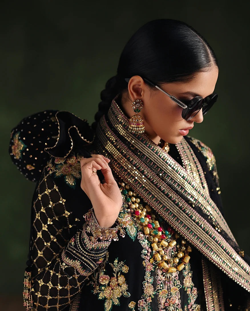 A woman wearing an ornate black embellished traditional outfit with heavy gold embroidery, a beaded shawl, large earrings, and black sunglasses, photographed against a dark green background.